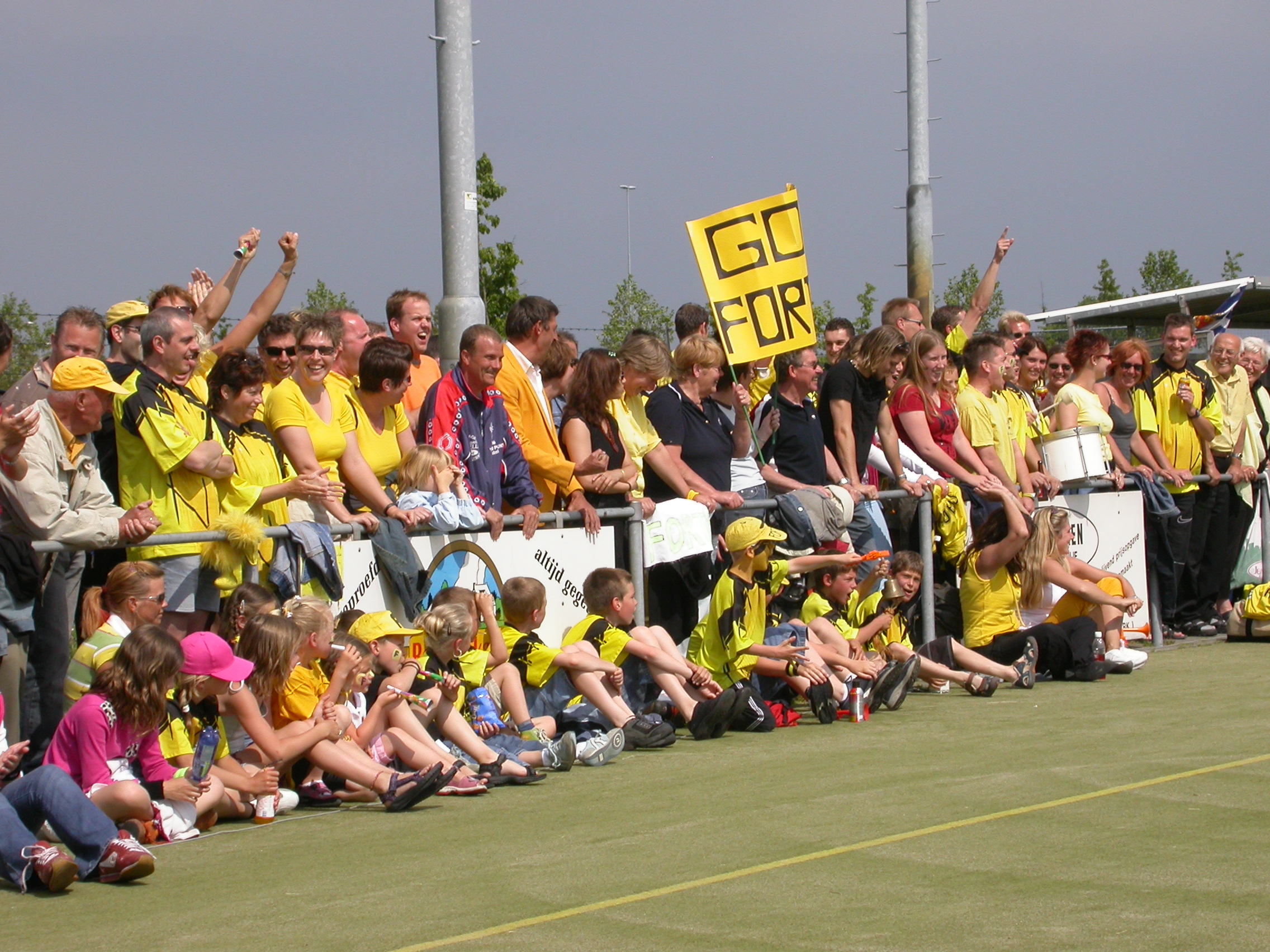 Supporters van Fortis staan langs het veld met geel-zwarte kleding en een spandoek.